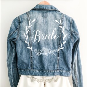 Könnte beinhalten: Eine blaue Jeansjacke mit einem weißen Blumenkranz und dem Wort "Bride" auf der Rückseite.