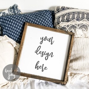 Peut inclure: Une pancarte en bois blanc avec un cadre brun, posée sur un lit de coussins blancs et bleus. La pancarte indique "your design here" en écriture cursive noire.