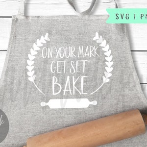 May include: A gray apron with the text "On your mark get set bake" in a white wreath of hearts. A rolling pin is lying on the apron.