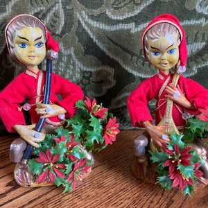 May include: Two vintage Christmas figurines. Each figure wears a red outfit and hat, holding a musical instrument. The figurines have painted faces with blue eyes and are surrounded by holly and poinsettias. The base is gold-colored.