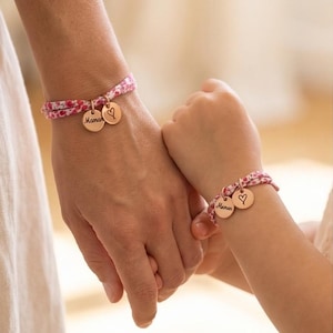 Op de afbeelding: Twee bijpassende armbanden met een roze en witte bloemenstoffen band. Elke armband bevat twee ronde bedels van roségoudkleurig metaal, waarvan er één is gegraveerd met het woord Maman en de andere een handgetekende hartvorm heeft.