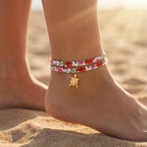 Può includere: Braccialetto alla caviglia con un motivo floreale rosso e rosa su sfondo bianco. Un ciondolo a forma di tartaruga dorata pende dal braccialetto. L'immagine mostra il braccialetto indossato su una caviglia su una spiaggia sabbiosa.