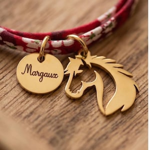 May include: A red floral fabric band featuring two gold-toned metal charms. One charm is a circular disc engraved with the name Margaux, and the other charm is a cutout silhouette of a horse head. Both charms are attached to the fabric band.