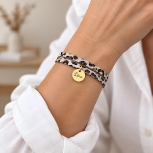 Peut inclure: Un bracelet à double tour avec un motif léopard dans des tons marron, noir et crème. Le bracelet comporte une petite breloque ronde en métal doré gravée du prénom Léna dans une police d'écriture cursive de couleur noire.