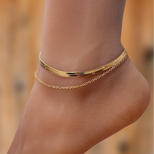 May include: A gold-coloured anklet featuring a herringbone pattern and a fine chain. The anklet is displayed on an ankle, highlighting its elegant and simple design. The jewellery is a stylish accessory.