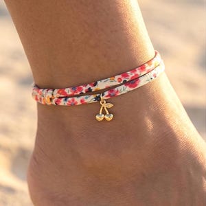 Op de afbeelding: Enkelbandje met bloemenprint en een gouden kersenbedel. Het enkelbandje heeft een witte basis met rode, roze en gele bloemen. De bedel is van goudkleurig metaal. Het enkelbandje wordt om de enkel gedragen.
