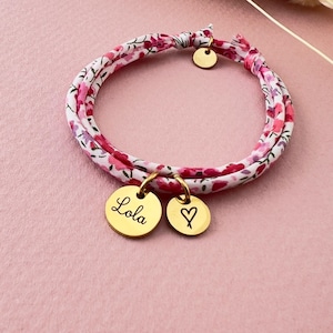 Peut inclure: Un bracelet en tissu à motifs floraux roses et rouges, avec des breloques dorées. L'une des breloques porte le nom "Lola" gravé, et l'autre un cœur. Le bracelet est en tissu blanc.