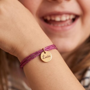 May include: A bracelet featuring a thin, shimmering magenta cord wrapped multiple times around the wrist, accented with a small, circular gold-toned metal charm engraved with the name Lucia in a dark, elegant script font.