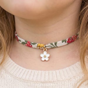 Peut inclure: Un collier ras-du-cou en tissu avec une base crème ornée d'un motif floral composé de fleurs rouges, de touches jaunes et de feuilles vertes. Le collier comporte une petite breloque en forme de fleur blanche à cinq pétales avec un centre doré.