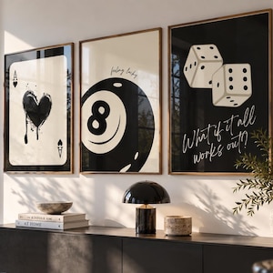 Pode incluir: Três quadros de parede emoldurados com uma carta de ás de copas em preto e branco, uma bola de bilhar número oito com o texto feeling lucky e dois dados brancos com o texto What if it all works out?, dispostos em uma linha horizontal.