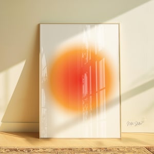 Peut inclure: Une impression d'art encadrée présentant un design abstrait. L'œuvre d'art met en valeur un dégradé vibrant de teintes orange et rouge, ressemblant à un soleil ou à une explosion de lumière circulaire, sur fond blanc doux. Le cadre est de couleur or clair.