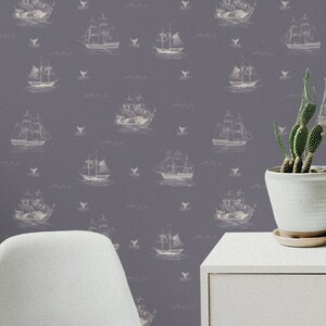 May include: Wallpaper with a repeating pattern of white vintage sailing ships on a muted purple backdrop. The design includes stylized waves and whale illustrations. A white cabinet, a cactus in a white pot, and a black speaker are in the foreground.