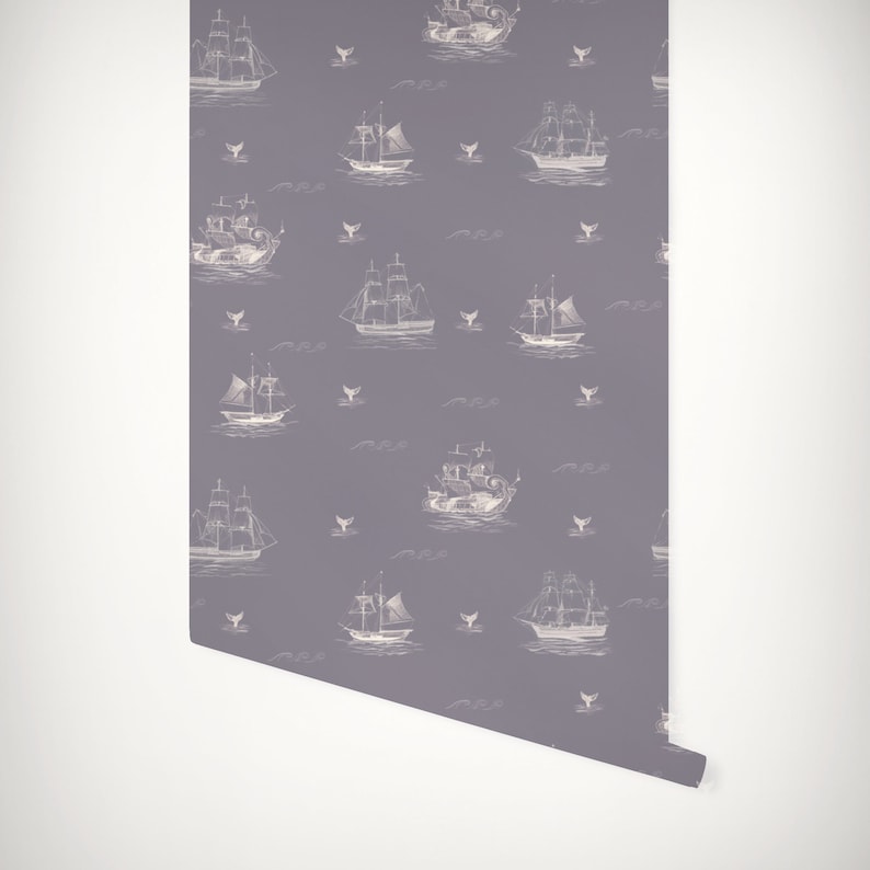 May include: Wallpaper roll with a repeating pattern of white sailing ships, whales, and waves on a grey background. The design has a nautical theme, suitable for a coastal or vintage interior. The roll is partially unrolled.
