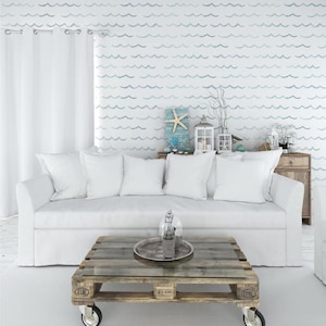 May include: A living room with a coastal theme. A white sofa with several white pillows sits in front of a wall with a wave pattern. A wooden coffee table with a glass top and a metal tray with a bottle and a glass sits in front of the sofa.