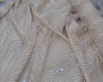 May include: Beige fabric with a delicate pattern of silver beaded feathers. The fabric has a subtle shimmer and is perfect for creating elegant garments or accessories.