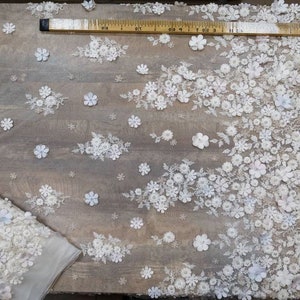 May include: White floral lace fabric with a textured, 3D effect. The fabric is shown on a wooden surface with a measuring tape.
