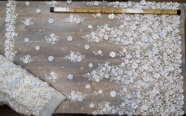 May include: White floral lace fabric with a textured, 3D effect. The fabric is shown on a wooden surface with a measuring tape.
