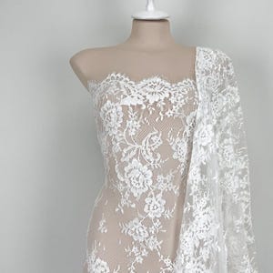 May include: White floral lace fabric draped over a mannequin. The lace has a delicate floral pattern with scalloped edges. The fabric is sheer, revealing the mannequin. Suitable for bridal or formal wear.