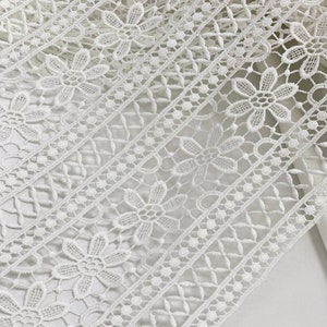 May include: White lace trim with a delicate floral pattern. The lace has a repeating design of small flowers and a geometric pattern.