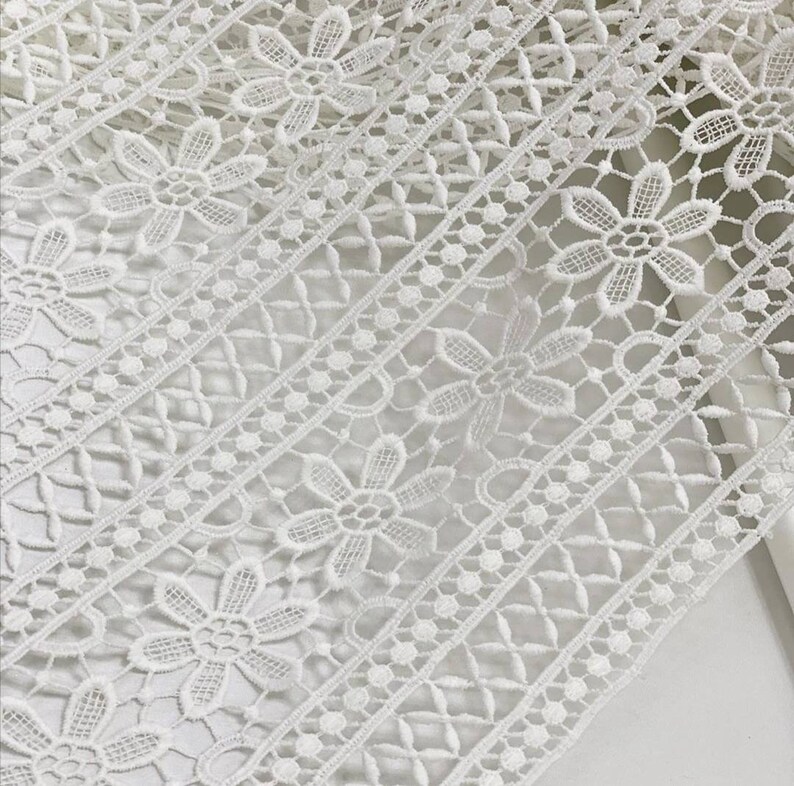 May include: White lace trim with a delicate floral pattern. The lace has a repeating design of small flowers and a geometric pattern.
