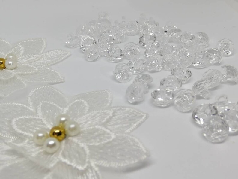 May include: Two white embroidered flower appliques featuring central clusters of four white pearls and one gold bead, accompanied by a collection of small, clear, faceted, round plastic buttons with a hole for sewing attachment.