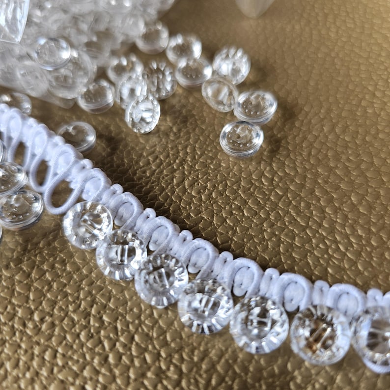 May include: A white loop trim featuring a row of small, clear, circular plastic buttons attached along the edge. The buttons are transparent with a faceted design, and the trim consists of a continuous series of white fabric loops.