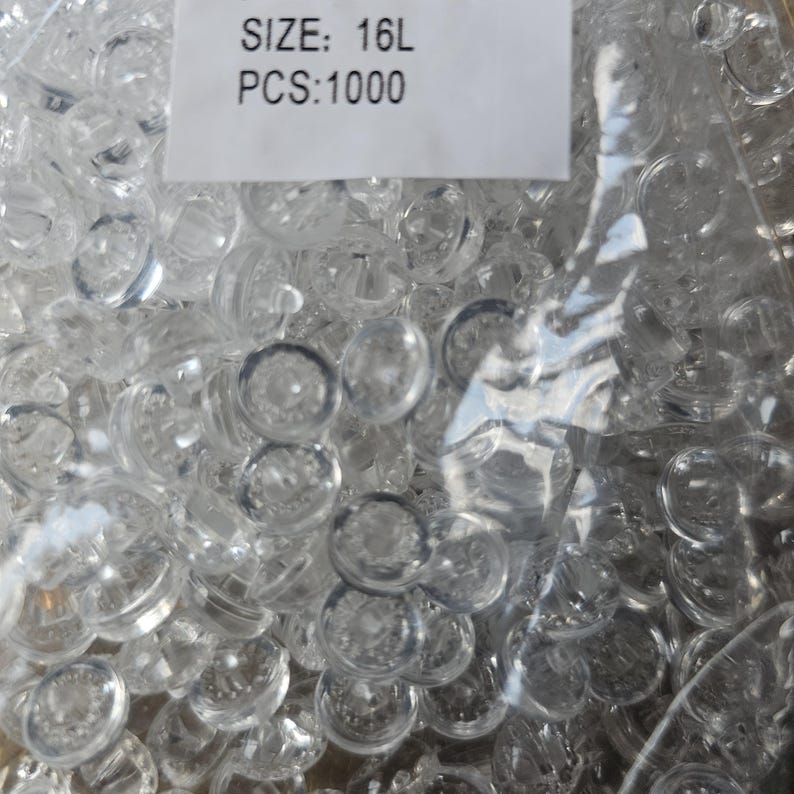 May include: A large collection of small, round, clear plastic buttons contained within a transparent plastic bag. A white label is attached to the top of the bag with the black printed text: SIZE: 16L PCS:1000. The buttons are translucent.