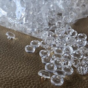 May include: A collection of small, round, clear plastic buttons with a shank back. The buttons are scattered across a surface, with many remaining inside a partially open, transparent plastic bag. Each button features a smooth, dome-shaped top.