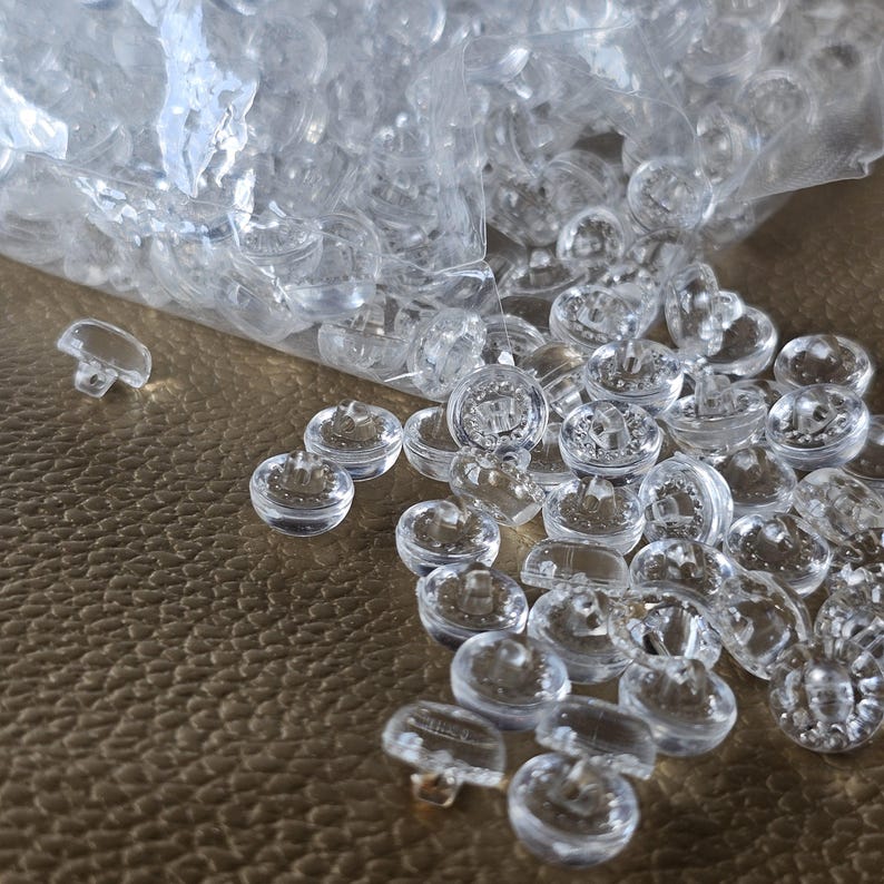 May include: A collection of small, round, clear plastic buttons with a shank back. The buttons are scattered across a surface, with many remaining inside a partially open, transparent plastic bag. Each button features a smooth, dome-shaped top.