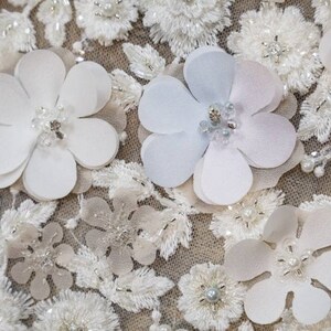 May include: Close-up of a white fabric with delicate floral embroidery and 3D flower embellishments. The flowers are made of white and light blue fabric and have a pearl-like center.
