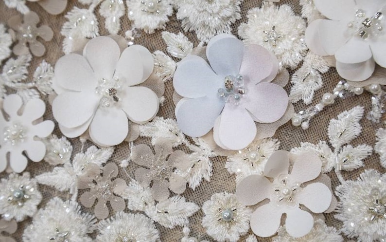May include: Close-up of a white fabric with delicate floral embroidery and 3D flower embellishments. The flowers are made of white and light blue fabric and have a pearl-like center.