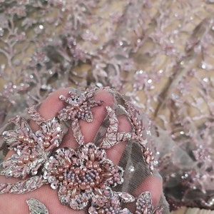 May include: Close-up of a delicate, beaded lace fabric with a floral pattern. The fabric is a light pink colour with silver accents and features a variety of beads and sequins.