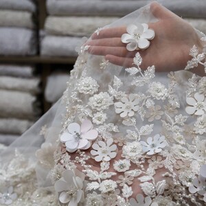 May include: White lace fabric with delicate floral embroidery and 3D flowers. The fabric is sheer and has a soft, romantic feel.