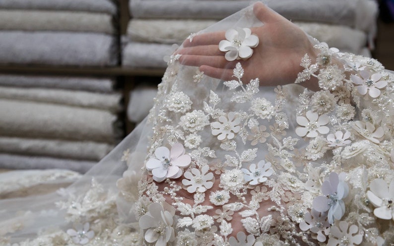 May include: White lace fabric with delicate floral embroidery and 3D flowers. The fabric is sheer and has a soft, romantic feel.