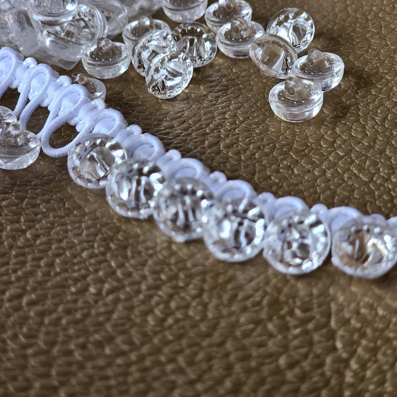 May include: A white elastic cord featuring a series of attached clear, round, faceted plastic buttons. Additional loose clear buttons are scattered nearby. The buttons have a smooth, reflective surface and a small center hole for attachment.