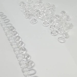 May include: A collection of clear, faceted, round buttons and a length of white button loop tape. The buttons are arranged in a loose cluster, while the tape features a series of evenly spaced, circular loops designed for fastening garments.
