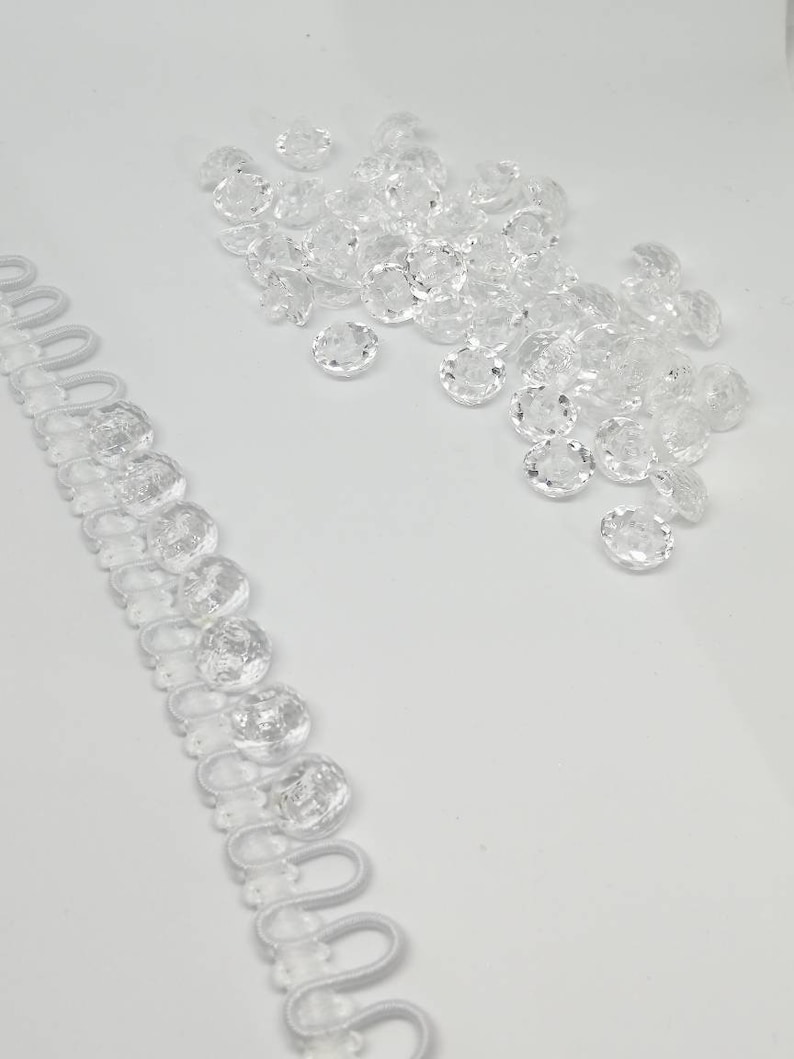 May include: A collection of clear, faceted, round buttons and a length of white button loop tape. The buttons are arranged in a loose cluster, while the tape features a series of evenly spaced, circular loops designed for fastening garments.