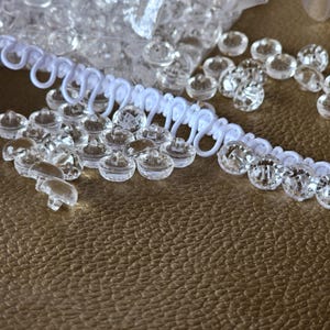 May include: A collection of small, clear, round plastic buttons with a faceted surface, accompanied by a length of white elastic loop trim. The buttons feature a shank on the underside for attachment, and the trim consists of a series of connected loops.
