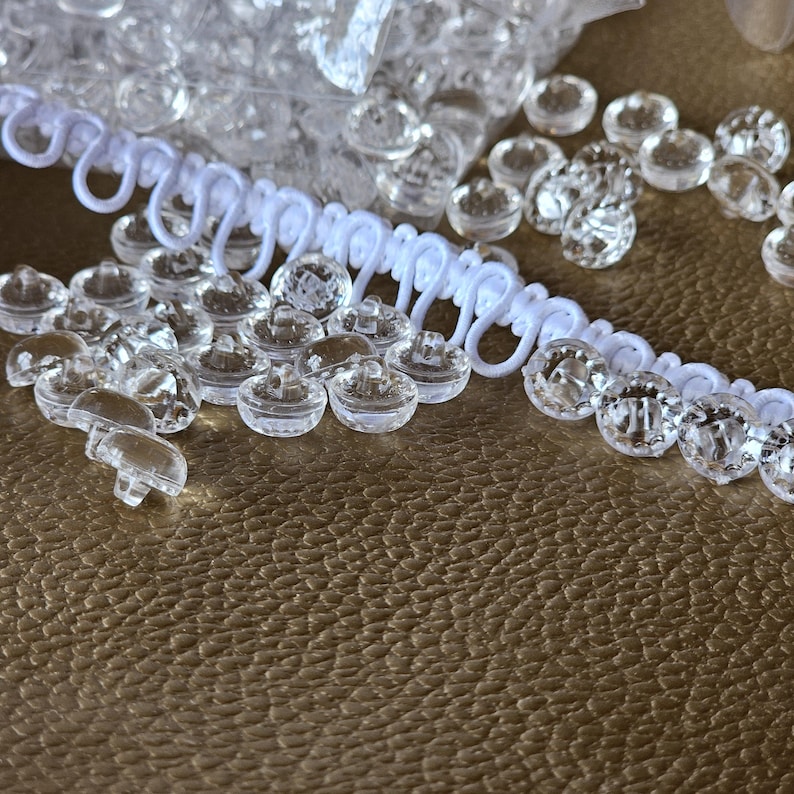 May include: A collection of small, clear, round plastic buttons with a faceted surface, accompanied by a length of white elastic loop trim. The buttons feature a shank on the underside for attachment, and the trim consists of a series of connected loops.