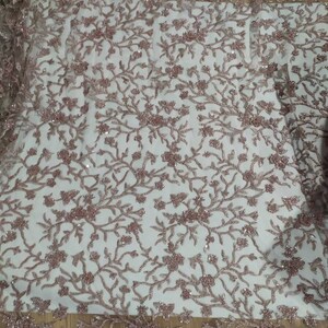 May include: A close-up of a delicate, rose-gold beaded lace fabric with a floral pattern. The fabric has a sheer, mesh-like background and is adorned with small, sparkling beads.