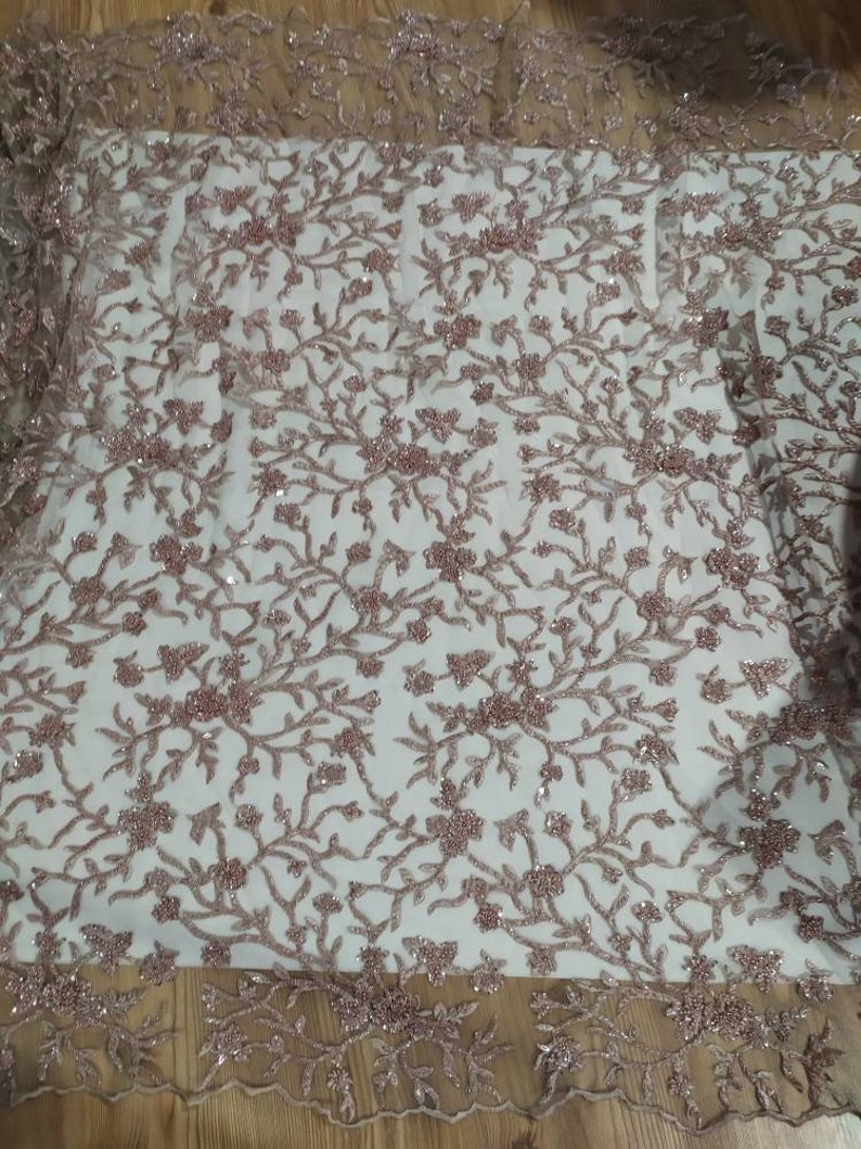 May include: A close-up of a delicate, rose-gold beaded lace fabric with a floral pattern. The fabric has a sheer, mesh-like background and is adorned with small, sparkling beads.