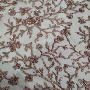 May include: A close-up of a delicate, white fabric with a floral pattern embroidered in a light pink colour. The fabric is covered in small, shiny beads that create a shimmering effect.