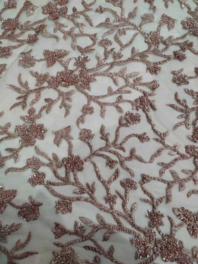 May include: A close-up of a delicate, white fabric with a floral pattern embroidered in a light pink colour. The fabric is covered in small, shiny beads that create a shimmering effect.