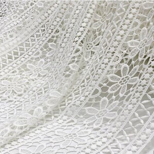 May include: White crochet lace fabric with a delicate floral pattern. The lace has a scalloped edge and is perfect for adding a touch of elegance to any project.
