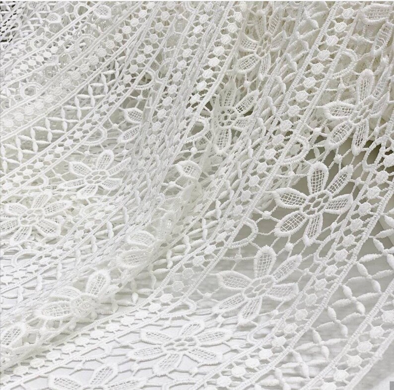 May include: White crochet lace fabric with a delicate floral pattern. The lace has a scalloped edge and is perfect for adding a touch of elegance to any project.