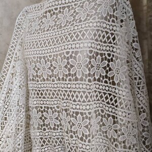 May include: White crochet fabric with a delicate floral pattern. The fabric has a geometric design with small flowers and a repeating pattern.