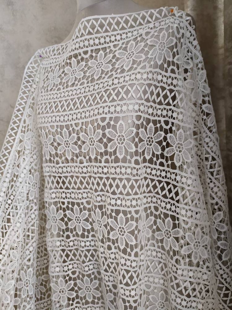 May include: White crochet fabric with a delicate floral pattern. The fabric has a geometric design with small flowers and a repeating pattern.