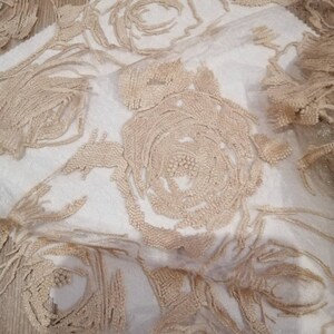 May include: Beige floral lace fabric with a delicate, embroidered pattern. The fabric is sheer and has a soft, flowing texture.