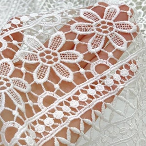 May include: White crochet lace fabric with a delicate floral pattern. The lace has a repeating design of small flowers and a larger flower with a center circle.