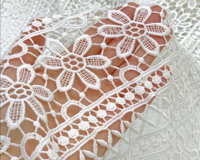May include: White crochet lace fabric with a delicate floral pattern. The lace has a repeating design of small flowers and a larger flower with a center circle.